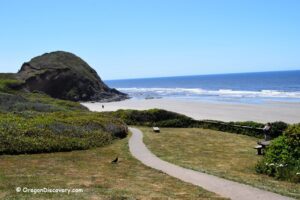 Ocean Beach Picnic Area (Ocean Beach Day Use Site) in Oregon: The picture "Ocean Beach grassy picnic overlook" features a grassy picnic area with benches and a paved path overlooking the sandy beach, with a rocky headland and gentle waves in the background.