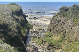 Tokatee Klootchman State Natural Site in Oregon: The picture "Tokatee Klootchman ravine with logs" shows a person standing on rocks next to a stream filled with driftwood, surrounded by steep, green cliffs and a forested hill in the distance.