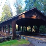 Cedar Crossing Bridge in Oregon: The picture "Cedar Crossing Bridge front view" displays a head-on view of the wooden covered bridge, with its peaked roof, large open entrance, and the words "Cedar Cross" visible above the entryway, surrounded by trees and a quiet country road.