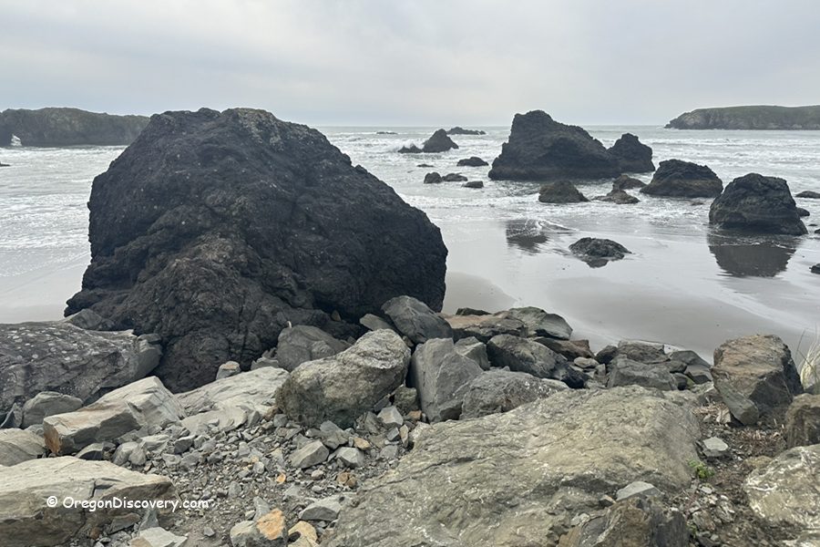 Myers Creek Beach in Oregon: The picture "Myers Creek Beach black rocks at low tide" displays large dark volcanic rocks scattered across the wet sand and shallow tidal pools, with waves breaking beyond and a cloudy sky overhead.