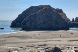 Myers Creek Beach in Oregon: The picture "Myers Creek Beach sea stacks and horseback rider" shows a wide sandy beach with a large rocky sea stack near the shoreline, a person riding a horse with two dogs walking alongside, and another person walking in the distance under a clear blue sky.