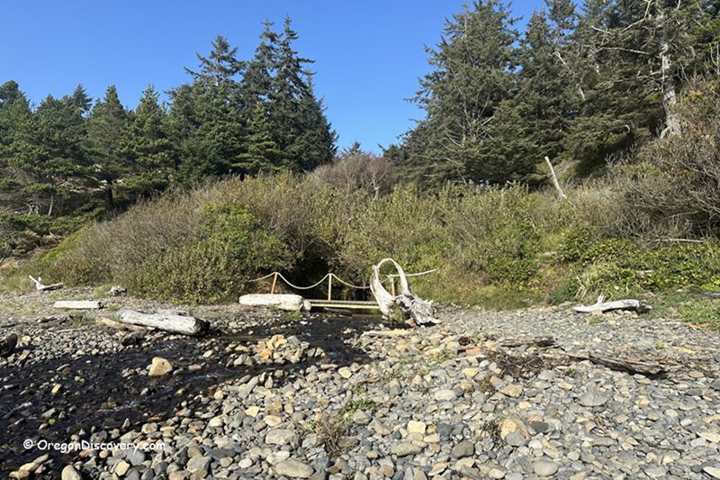 Happy Beach in Oregon: The picture "Happy Beach in Oregon – creek with driftwood and rocky shore" shows a small creek flowing through a rocky area toward the beach, with large pieces of driftwood and dense green bushes and pine trees under a bright blue sky.