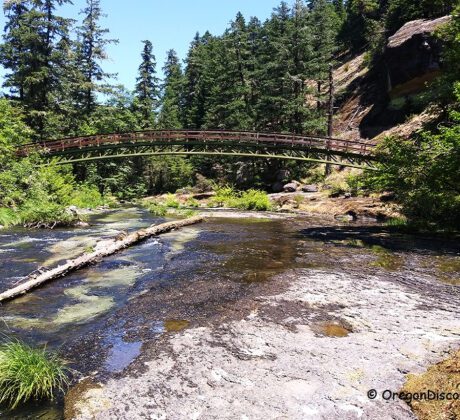 Wolf Creek Falls Recreation Area in Oregon: The picture "Wolf Creek Falls Recreation Area in Oregon – footbridge over creek and forest" displays a footbridge arching over a flowing creek, surrounded by green forest and rocky banks, with a large rock formation in the background.