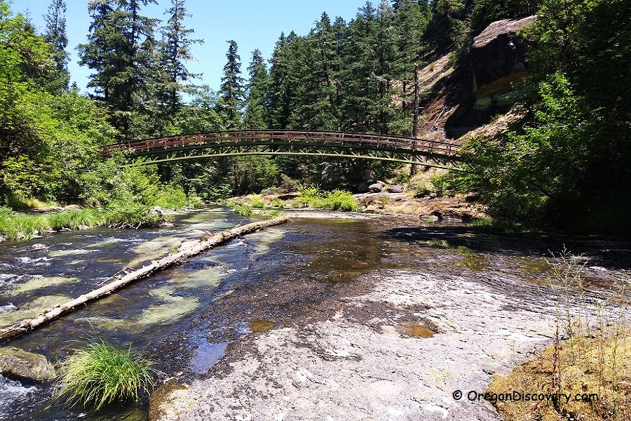 Wolf Creek Recreation Area Wolf Creek Falls Recreation Area in Oregon: The picture "Wolf Creek Falls Recreation Area in Oregon – footbridge over creek and forest" displays a footbridge arching over a flowing creek, surrounded by green forest and rocky banks, with a large rock formation in the background.