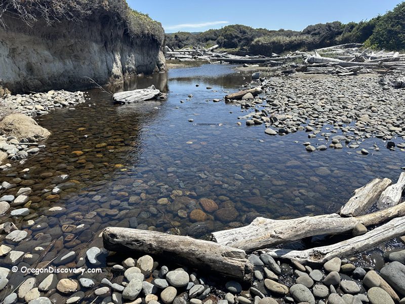 Muriel O. Ponsler Memorial State Scenic Viewpoint in Oregon: The picture "Muriel O. Ponsler Memorial State Scenic Viewpoint creek, pebbles, and driftwood" shows a shallow, clear creek flowing over smooth pebbles with weathered driftwood scattered along the banks and a sandy bluff in the background, beneath a sunny sky.