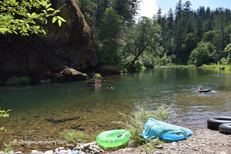 Dumont Creek Campground in Oregon: The picture "Dumont Creek Campground in Oregon – swimming and inner tubes in scenic river bend" features people swimming and floating on inner tubes in a calm bend of a river, surrounded by rocky banks, green forest, and tall trees, with inflatable tubes and towels on the shore.