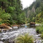 Dumont Creek Campground in Oregon: The picture "Dumont Creek Campground in Oregon – river with mossy rocks and dense forest backdrop" shows a flowing river bordered by moss-covered boulders and thick vegetation, with evergreen trees rising up steep hillsides and a blue inner tube floating in the water.