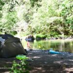 White Creek Campground in Oregon: The picture "White Creek Campground in Oregon – tent and camping gear by a wooded creek" shows a large tent set up on a gravelly bank beside a calm, tree-lined creek, with colorful camping chairs, float tubes, and a kayak scattered around under leafy green trees.