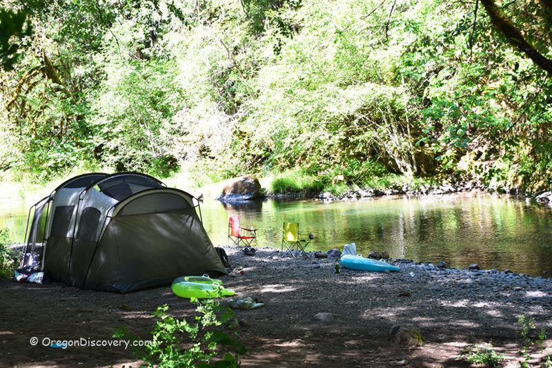White Creek Campground in Oregon: The picture "White Creek Campground in Oregon – tent and camping gear by a wooded creek" shows a large tent set up on a gravelly bank beside a calm, tree-lined creek, with colorful camping chairs, float tubes, and a kayak scattered around under leafy green trees.