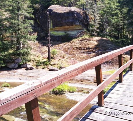 Wolf Creek Falls Recreation Area in Oregon: The picture "Wolf Creek Falls Recreation Area in Oregon – footbridge over creek and forest" displays a footbridge arching over a flowing creek, surrounded by green forest and rocky banks, with a large rock formation in the background.