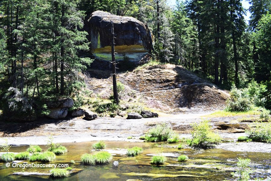 Wolf Creek Falls Recreation Area in Oregon: The picture "Wolf Creek Falls Recreation Area in Oregon – large rock and forested hillside" features a massive boulder sitting on a forested hillside above a creek, with patches of sunlight and tall evergreen trees in the background.
