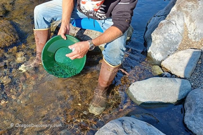 Quartzville Creek - Recreational Gold Panning and Rockhounding in Oregon: The picture "Quartzville Creek - panning for gold at the creek" shows a person wearing boots and jeans kneeling in shallow water, using a green gold pan to sift for gold among rocks and gravel by the stream.