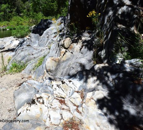 South Umpqua River Rockhounding in Oregon: The picture "South Umpqua River Rockhounding in Oregon – gray rock outcrops along riverside trail" shows smooth, light and dark gray rock formations covered in patches of grass and wildflowers, with a person walking along a sandy trail beside the river in the background.