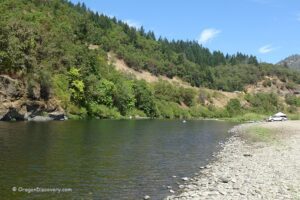 Myrtle Creek Rockhounding in Oregon: The picture "Myrtle Creek Rockhounding in Oregon – riverbank with parked car and tree-lined hillside" shows a tranquil bend of the river with a stony shore in the foreground, a white car parked near the water's edge, and lush, forested hills forming a green backdrop beneath a blue sky.