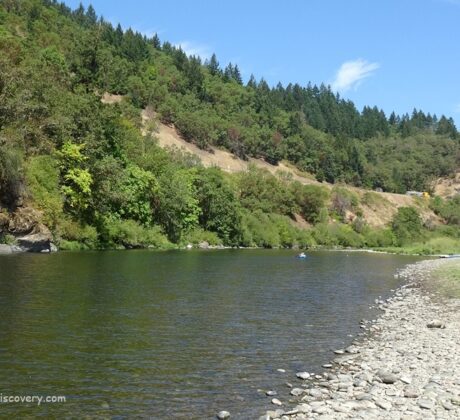 Myrtle Creek Rockhounding in Oregon: The picture "Myrtle Creek Rockhounding in Oregon – riverbank with parked car and tree-lined hillside" shows a tranquil bend of the river with a stony shore in the foreground, a white car parked near the water's edge, and lush, forested hills forming a green backdrop beneath a blue sky.