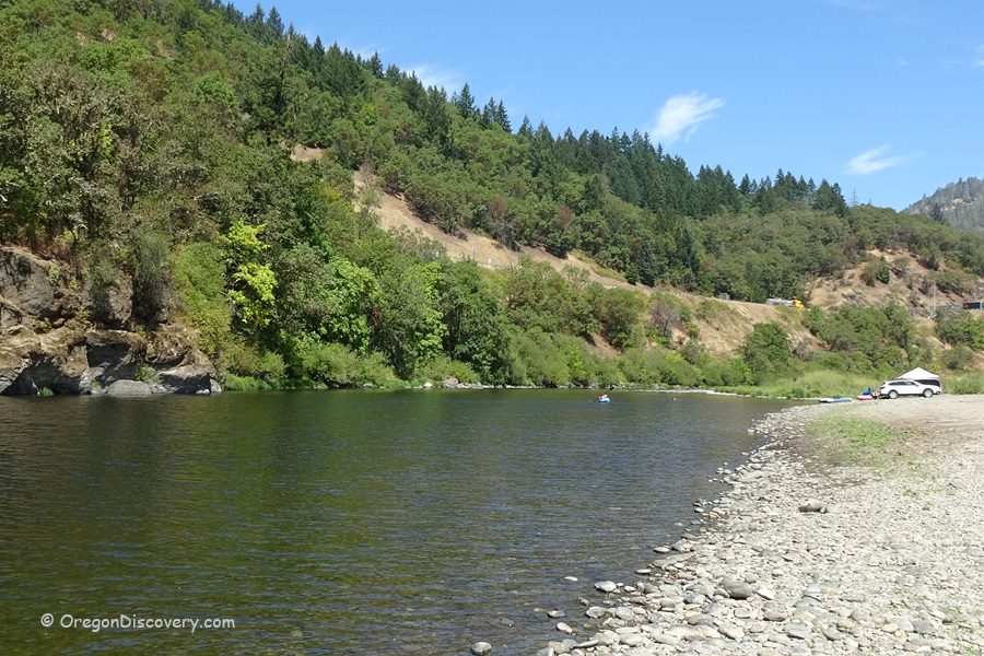Myrtle Creek Rockhounding in Oregon: The picture "Myrtle Creek Rockhounding in Oregon – riverbank with parked car and tree-lined hillside" shows a tranquil bend of the river with a stony shore in the foreground, a white car parked near the water's edge, and lush, forested hills forming a green backdrop beneath a blue sky.