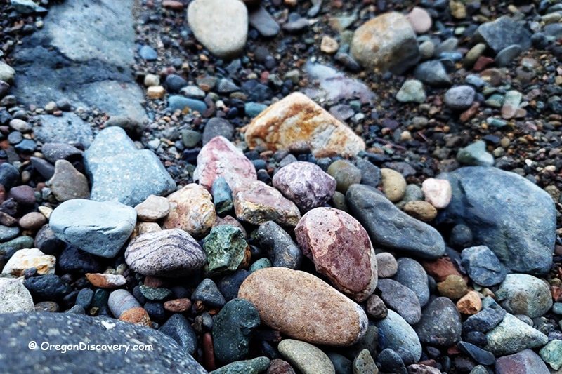 Quartzville Creek - Recreational Gold Panning and Rockhounding in Oregon: The picture "Quartzville Creek - colorful river rocks" features a close-up of various rounded and jagged river rocks in shades of pink, tan, gray, and green scattered along the gravelly creek shore.