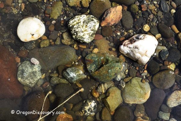 Myrtle Creek Rockhounding in Oregon: The picture "Myrtle Creek Rockhounding in Oregon – close-up of quartz and green rocks underwater" displays a variety of stones, including white, green, and brown rocks submerged in clear, shallow water, revealing their natural textures and colors.