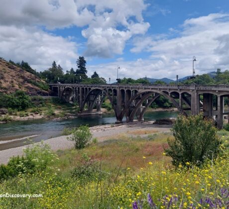 Myrtle Creek Rockhounding in Oregon: The picture "Myrtle Creek Rockhounding in Oregon – historic arched bridge over the river" shows a classic concrete arch bridge crossing the green river, surrounded by wildflowers and shrubs in the foreground, with tree-covered hills and partly cloudy skies in the background.