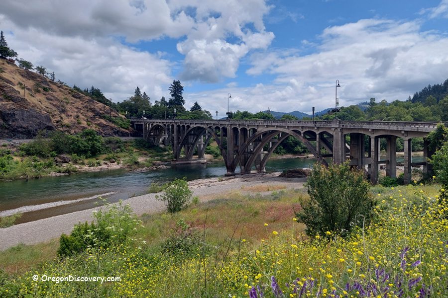 Myrtle Creek Rockhounding in Oregon: The picture "Myrtle Creek Rockhounding in Oregon – historic arched bridge over the river" shows a classic concrete arch bridge crossing the green river, surrounded by wildflowers and shrubs in the foreground, with tree-covered hills and partly cloudy skies in the background.