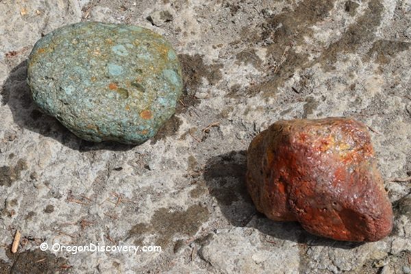 Ash Flat Campground in Oregon: The picture "Ash Flat Campground in Oregon – close-up of green and red stones on riverbed" features a close-up of two large, rounded rocks, one greenish and one reddish, lying on a dry, textured stone surface beside the river.