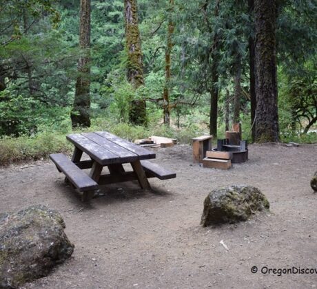Boulder Creek Campground in Oregon: The picture "Boulder Creek Campground in Oregon – picnic table and fire ring in the forest" shows a wooden picnic table and benches sitting on bare earth surrounded by large mossy boulders, with a metal fire ring and wooden seats nearby, all set in a dense, green forest.