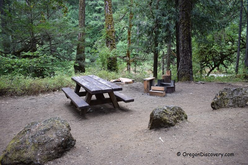 Boulder Creek Campground in Oregon: The picture "Boulder Creek Campground in Oregon – picnic table and fire ring in the forest" shows a wooden picnic table and benches sitting on bare earth surrounded by large mossy boulders, with a metal fire ring and wooden seats nearby, all set in a dense, green forest.