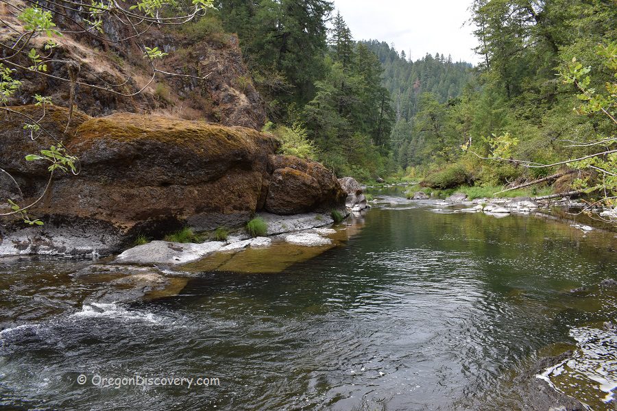 Ash Flat Campground in Oregon: The picture "Ash Flat Campground in Oregon – mossy rock cliffs and forested river bend" shows a scenic river curve with clear water, bordered by large moss-covered rock cliffs on the left and lush green forested hills in the background.