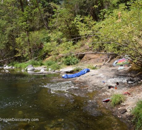 Ash Flat Campground in Oregon: The picture "Ash Flat Campground in Oregon – rocky shore with gear and floating tubes" depicts a rocky riverbank lined with green bushes, where a blue sleeping bag and various personal items are scattered near the water's edge, along with black inflatable tubes ready for floating.