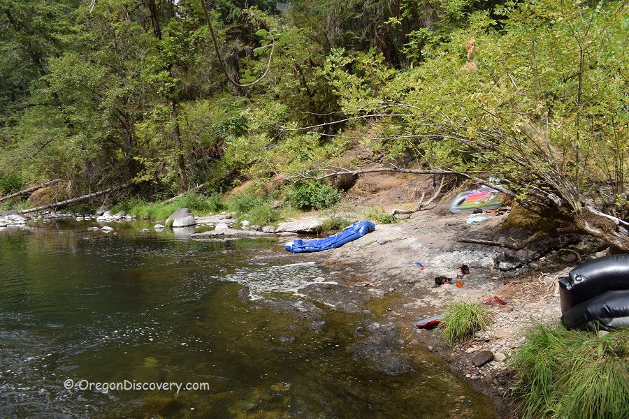 Ash Flat Campground in Oregon: The picture "Ash Flat Campground in Oregon – rocky shore with gear and floating tubes" depicts a rocky riverbank lined with green bushes, where a blue sleeping bag and various personal items are scattered near the water's edge, along with black inflatable tubes ready for floating.