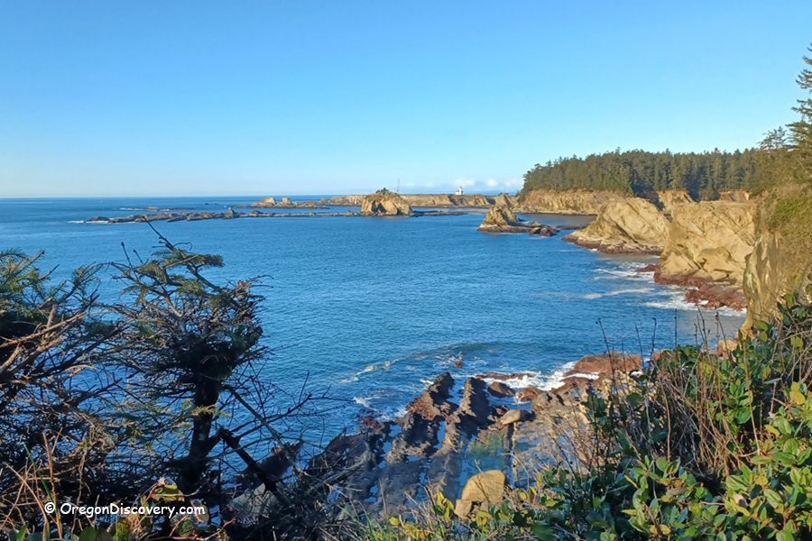 Cape Arago Lighthouse in Oregon: The picture "Cape Arago Lighthouse distant view across the bay" features a sweeping view of a deep blue bay lined with rocky cliffs and evergreen trees, with the Cape Arago Lighthouse visible in the distance atop a headland under a clear blue sky.