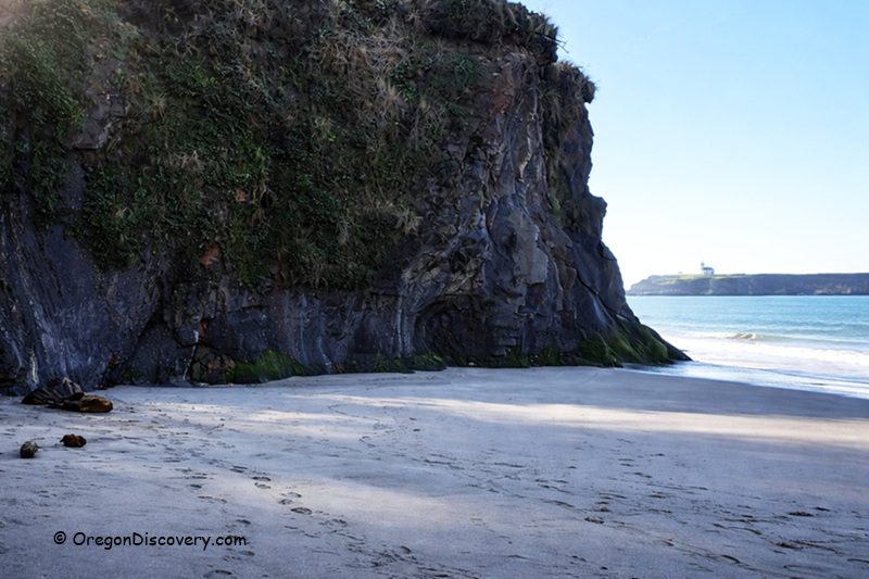 Lighthouse Beach in Oregon: The picture "Lighthouse Beach cliff and distant lighthouse" displays a sandy cove bordered by a steep, moss-covered rock cliff on the left, with the ocean and a distant lighthouse visible on the horizon.