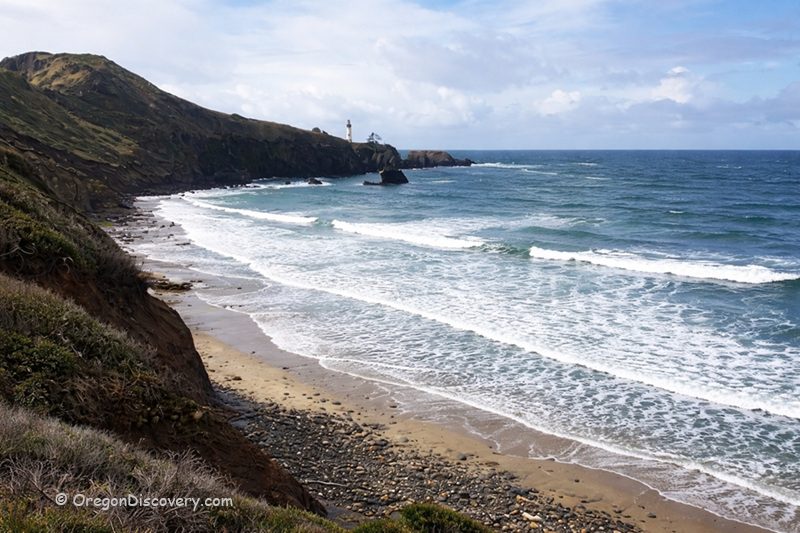 Starfish Cove in Oregon: The picture "Starfish Cove sandy beach and lighthouse" features a sweeping view of a sandy and pebbly beach curving along the coast, with white waves crashing onshore and a tall lighthouse visible on the distant headland under a partly cloudy sky.