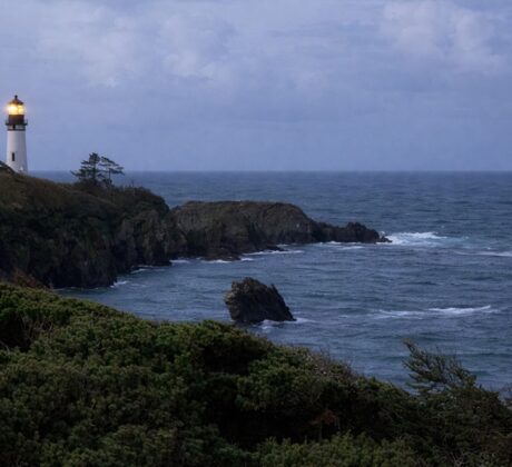 Starfish Cove in Oregon: The picture "Starfish Cove lighthouse at dusk" captures a moody scene of the lighthouse shining its light atop a dramatic seaside cliff, surrounded by dark green foliage and choppy ocean waters as dusk settles under a cloudy sky.