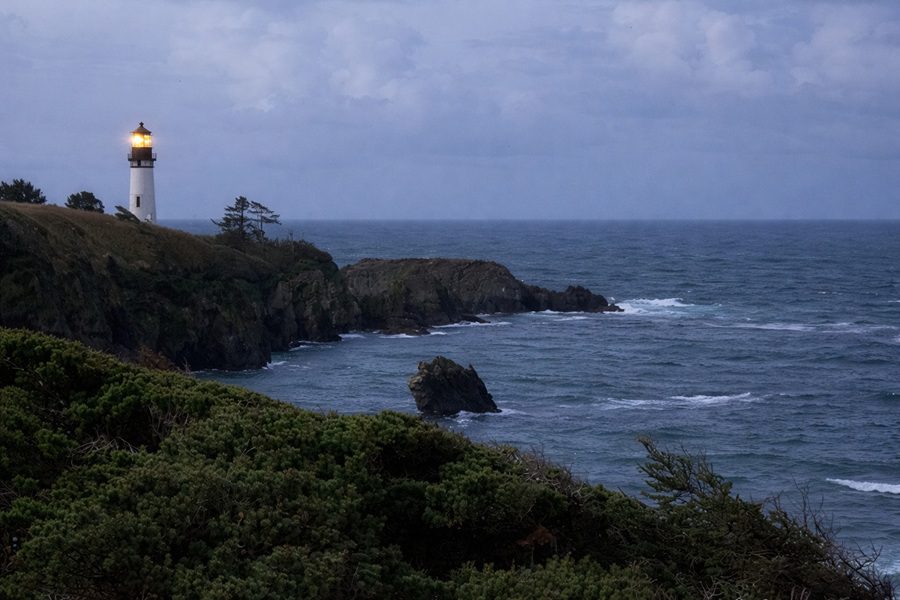 Starfish Cove in Oregon: The picture "Starfish Cove lighthouse at dusk" captures a moody scene of the lighthouse shining its light atop a dramatic seaside cliff, surrounded by dark green foliage and choppy ocean waters as dusk settles under a cloudy sky.