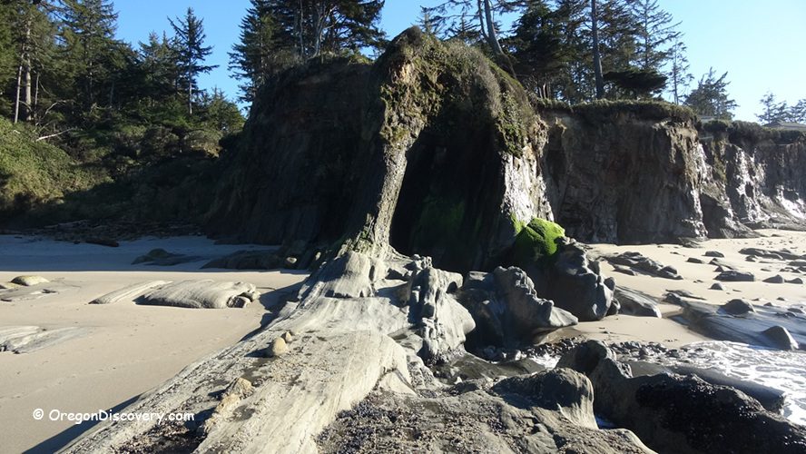 Lighthouse Beach in Oregon: The picture "Lighthouse Beach unique rock formation" features a dramatic, eroded rock formation jutting out from the sand and cliffs, partially covered with bright green moss, with the beach and forest backdrop under a sunny sky.