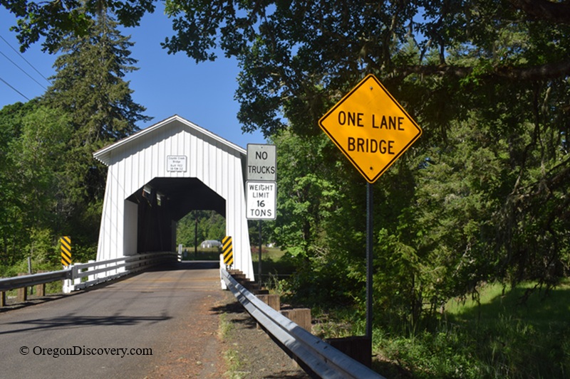 Coyote Covered Bridge in Oregon: The picture "one lane bridge entrance with signs" shows a white covered bridge at the entrance to Coyote Covered Bridge, with a yellow diamond-shaped sign that reads "ONE LANE BRIDGE" and other signs indicating no trucks and a weight limit, surrounded by trees and a paved road. 