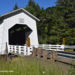 Coyote Covered Bridge in Oregon: The picture "side view of covered bridge" captures the exterior side angle of the white Coyote Creek Covered Bridge, with wooden fencing, metal guard rails, and green forested hillsides in the background.