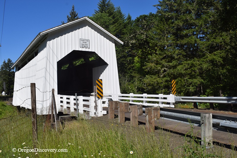 Coyote Covered Bridge in Oregon: The picture "side view of covered bridge" captures the exterior side angle of the white Coyote Creek Covered Bridge, with wooden fencing, metal guard rails, and green forested hillsides in the background.
