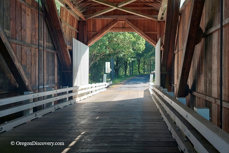 Coyote Covered Bridge in Oregon: The picture "inside view of covered bridge" shows the interior of Coyote Covered Bridge with wooden walls, beams, and planks, and sunlight streaming in from the exit, framing a tree-lined road outside.