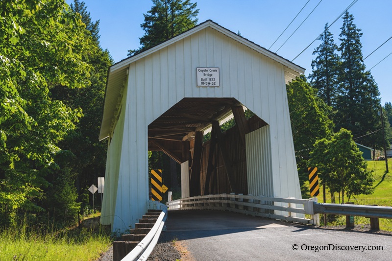 Coyote Covered Bridge in Oregon: The picture "Coyote Creek Bridge entrance" features the front view of the historic white wooden Coyote Creek Covered Bridge, built in 1922, with sunlit trees and a curving road leading into the structure.
