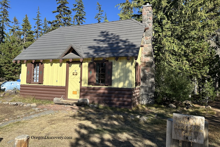 Olallie Lake in Oregon: The picture "Olallie Lake Guard Station Cabin" features a small, rustic cabin with yellow and brown painted walls, shuttered windows, and a stone chimney, set among tall evergreen trees with a wooden sign in front.