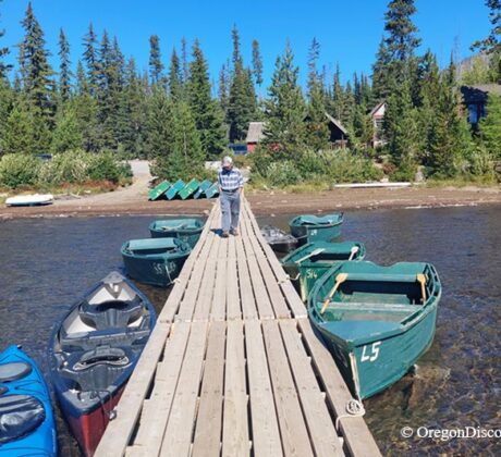 Olallie Lake in Oregon: The picture "dock with boats at shore" shows a wooden dock leading out into shallow water, with green rowboats and blue canoes tied up on both sides, and a person walking down the dock toward cabins and pine trees on the shore.
