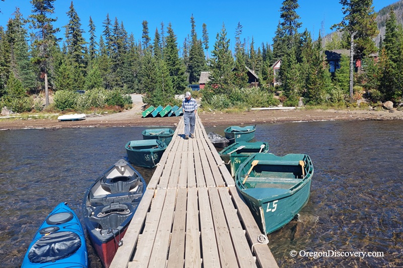 Olallie Lake in Oregon: The picture "dock with boats at shore" shows a wooden dock leading out into shallow water, with green rowboats and blue canoes tied up on both sides, and a person walking down the dock toward cabins and pine trees on the shore.