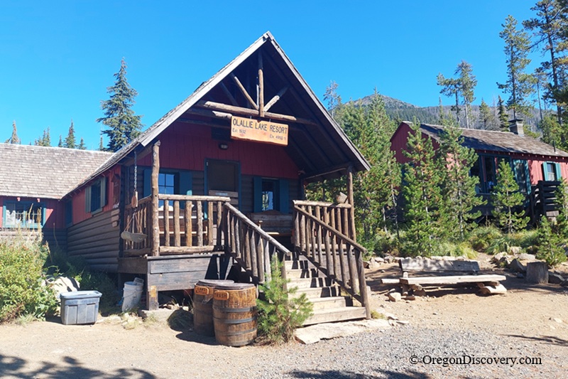 Olallie Lake in Oregon: The picture "Olallie Lake Resort Office" shows a log cabin building with a peaked roof, wooden railings, and a sign reading 'Olallie Lake Resort' above the porch, with scattered barrels and other wooden cabins surrounded by pine trees.
