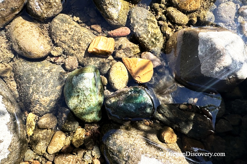 North Santiam River Rockhounding in Oregon: The picture "green jasper and river stones" shows a close-up of smooth river rocks underwater, including shiny green jasper stones and pieces of orange and brown rock.