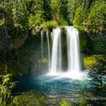 Koosah Falls in Oregon: The picture "Koosah Falls waterfall wide view" shows a stunning, double-plume waterfall pouring over a mossy cliff into a deep blue pool, surrounded by lush green forest and bright sunlight.