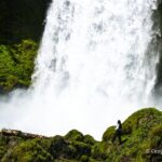 Sahalie Falls in Oregon: The picture "Sahalie Falls up close with person for scale" shows a powerful white waterfall crashing down a mossy green cliff, while a person stands on rocks near the misty base, highlighting the impressive size of the falls.