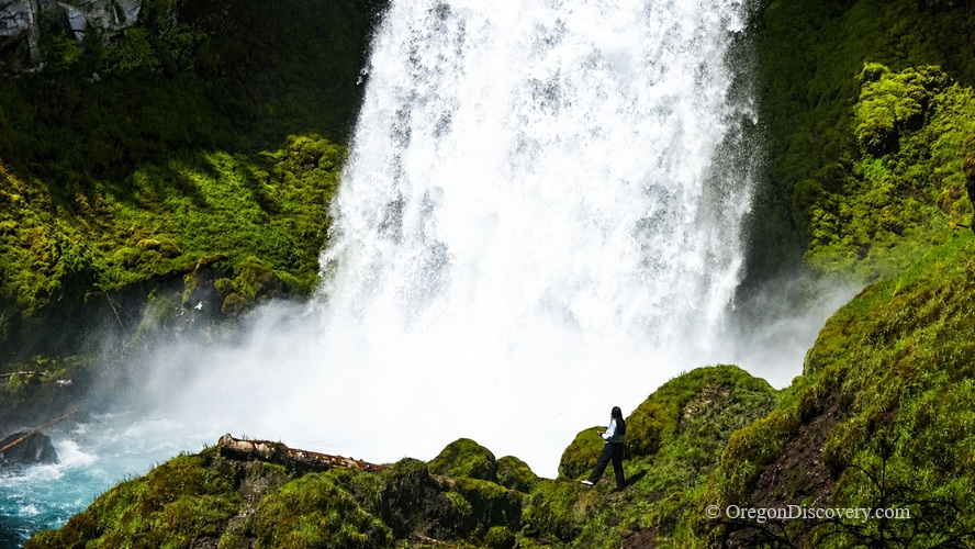 Sahalie Falls in Oregon: The picture "Sahalie Falls up close with person for scale" shows a powerful white waterfall crashing down a mossy green cliff, while a person stands on rocks near the misty base, highlighting the impressive size of the falls.