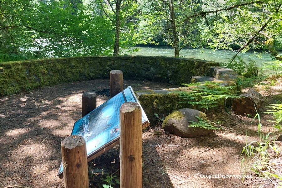 McKenzie River Trail in Oregon: The picture "McKenzie River Trail interpretive sign by riverside" shows an informational sign set into the ground beside a mossy stone wall, with trees and the flowing McKenzie River in the background.